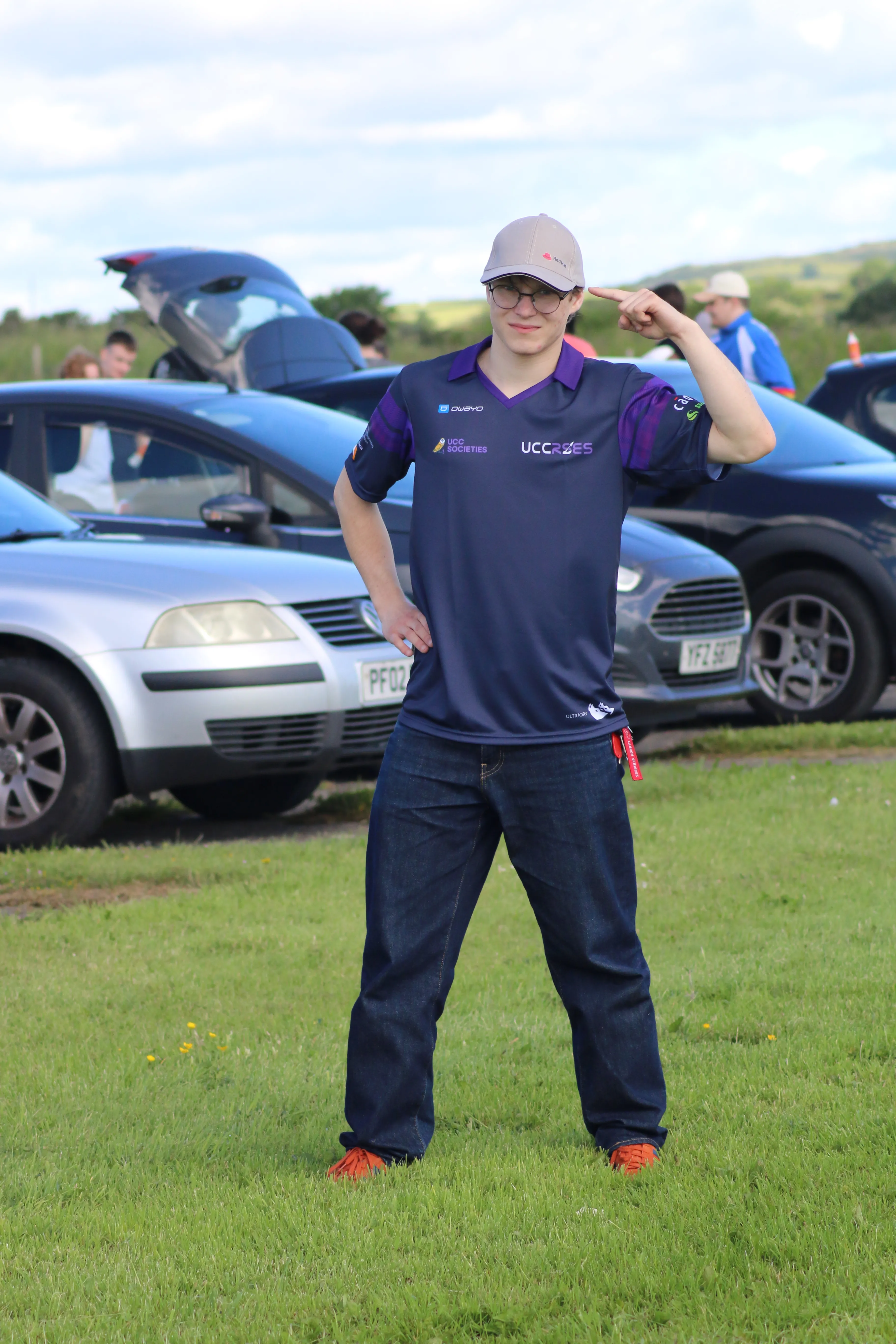 Standing outdoors on a bright day, smiling and pointing at a cap